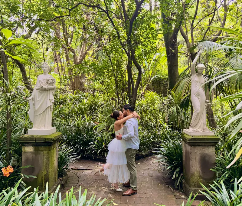 Karyme y Josué se besan en el sendero de ladrillo del jardín, entre dos estatuas de mármol